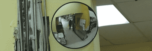 Convex mirror showing my small full-bodied reflection clad in black, through a hallway. On the left and right sides of the mirror are cables/scaffolding and a partially obscured light panel respectively.